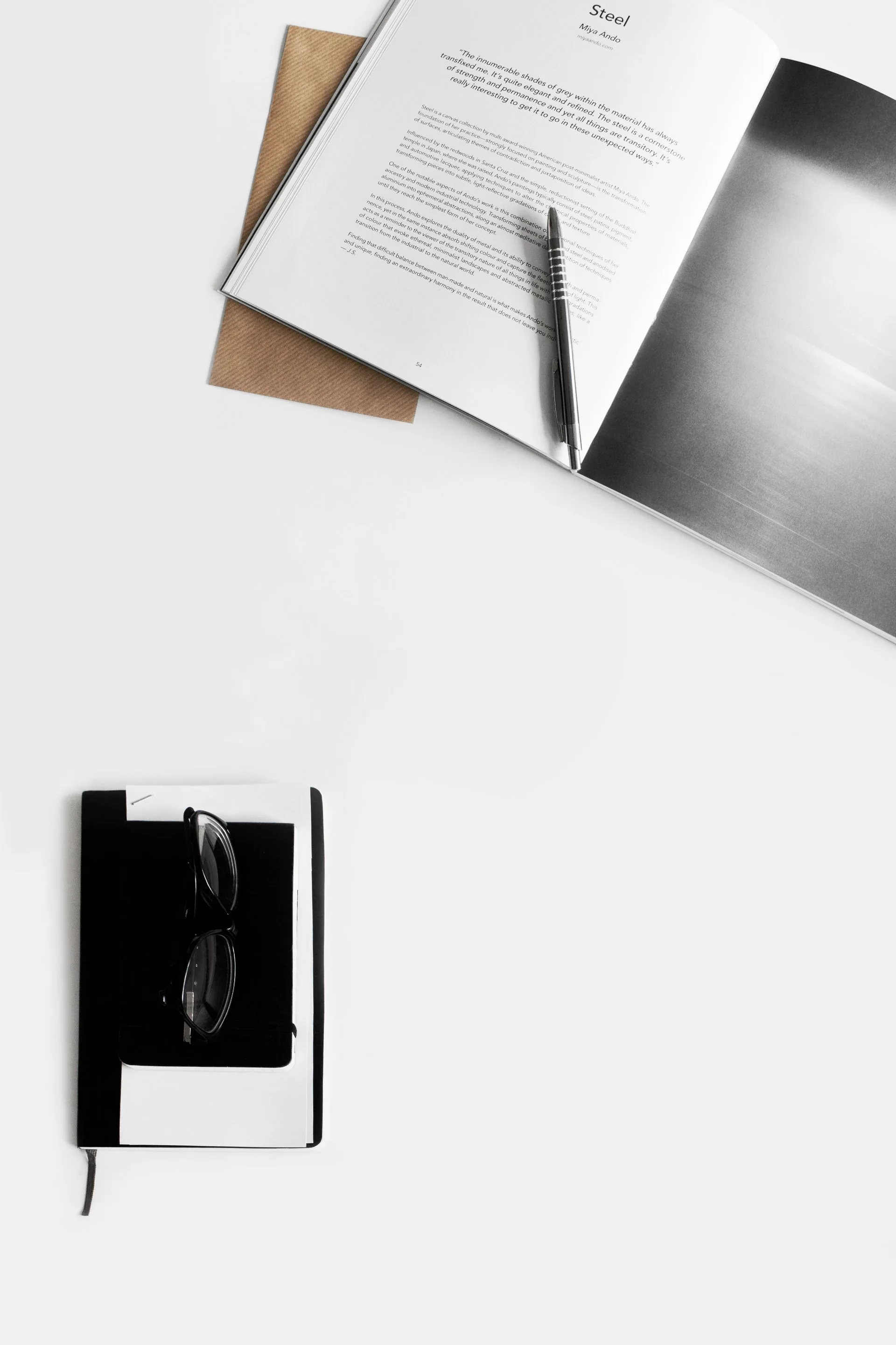 Livro aberto, caneta, óculos e caderno em uma mesa clara.