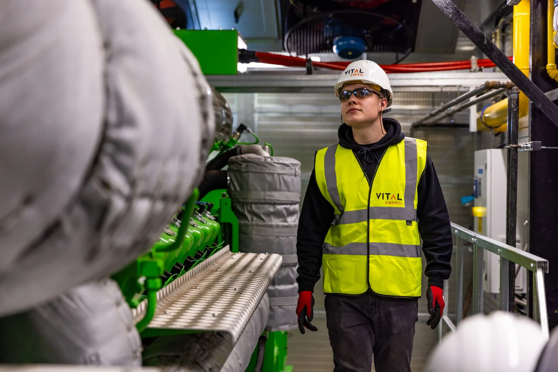 Profissional com capacete, óculos e colete observa equipamentos industriais.